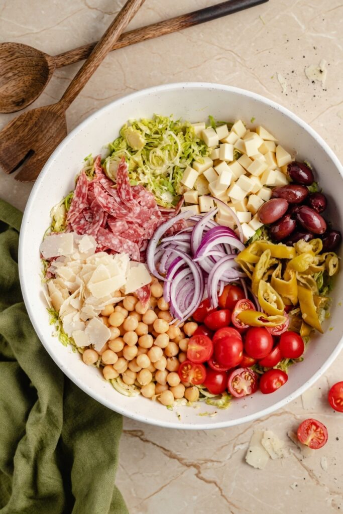 Ingredients for Italian Chopped Brussels Sprouts Salad including brussels sprouts chickpeas salami olives provolone and pepperoncini on marble surface
