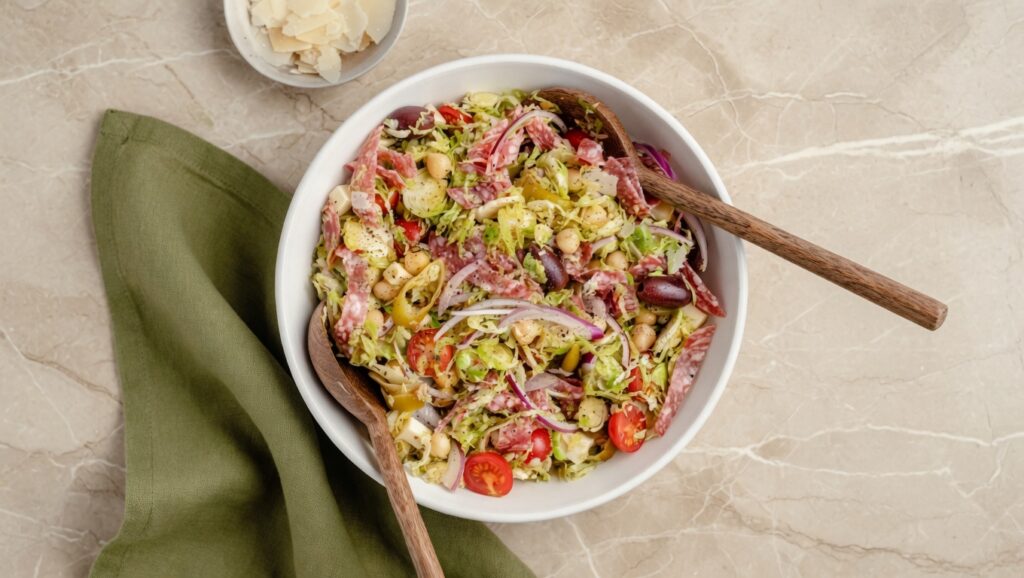 Italian Chopped Brussels Sprouts Salad in a large white bowl with salami olives chickpeas provolone and pepperoncini on a wooden table