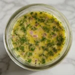 Lemon dill vinaigrette in a glass jar with fresh dill and lemon slices on a white marble surface